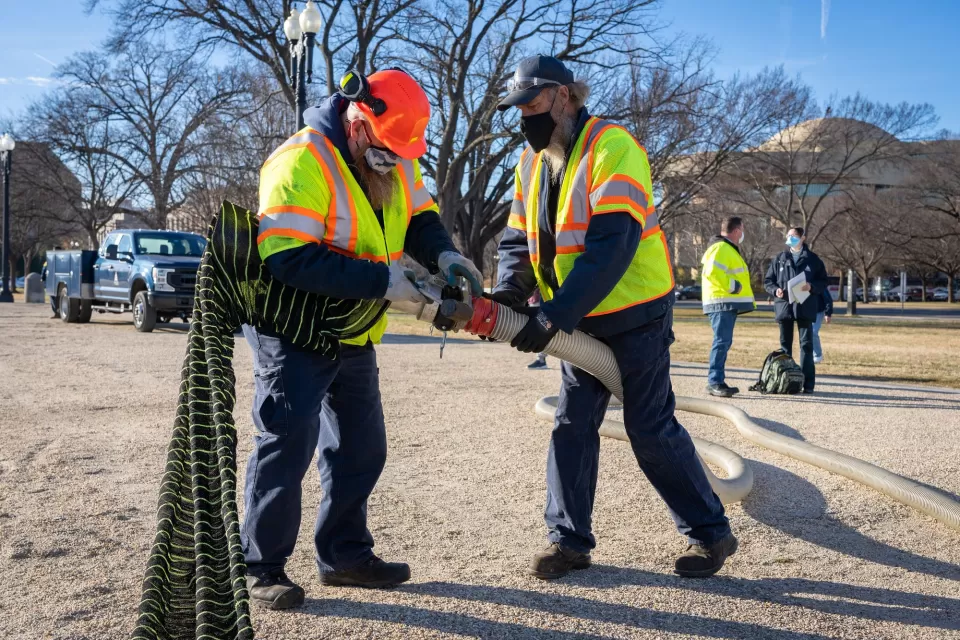 Frank Bussler (left), Engineering Equipment Operator for Capitol Grounds, helps Richard Caselman, Heavy Mobile Equipment Mechanic Assistant Supervisor for Capitol Grounds, attach silt sock material to the end of a hose during a demonstration of the jurisdiction's new mulch blower. 